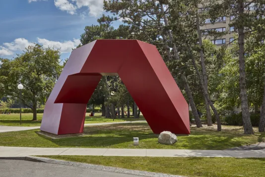 Red painted steel geometric sculpture with hard edges and rectangular shapes within on a small grass field. 