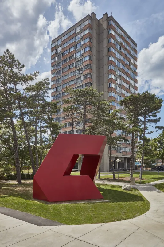 Red painted steel geometric sculpture with hard edges and rectangular shapes within on a small grass field with a tall dorm building in the background.