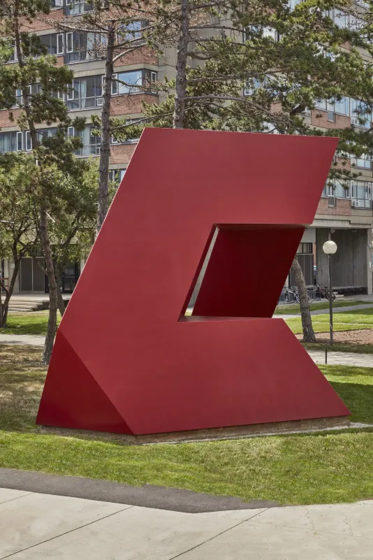 Red painted steel geometric sculpture with hard edges and rectangular shapes within on a small grass field. 