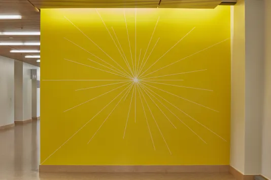 Bright yellow wall with a drawing featuring thin lines that come out from the center of the wall to form a sun like shape. 
