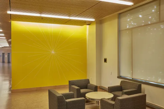 Bright yellow wall with a drawing featuring thin lines that come out from the center of the wall to form a sun like shape.