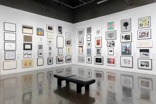 Installation view with framed artworks lining the walls floor to ceiling and a leather bench in the center of the gallery.
