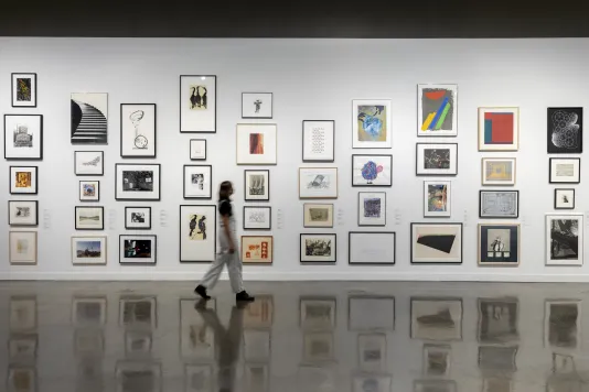 Installation view with framed artworks lining the wall and person in white overalls in motion walking through gallery.