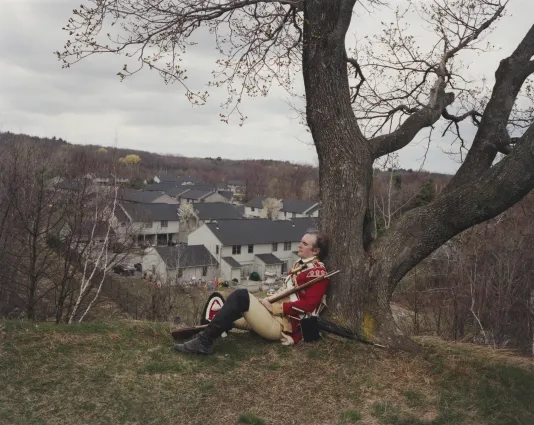 Photograph of a British Redcoat re-enactor sitting under a tree on some grass overlooking a suburb. 