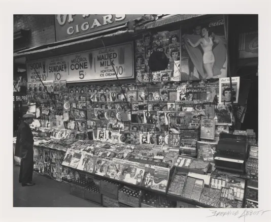 Black and white photograph of a newsstand full of newspapers and magazines with a person growing on the left.