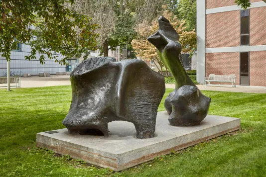 Large-scale bronze abstract sculpture of a figure on a stone pedestal in a grassy courtyard. 