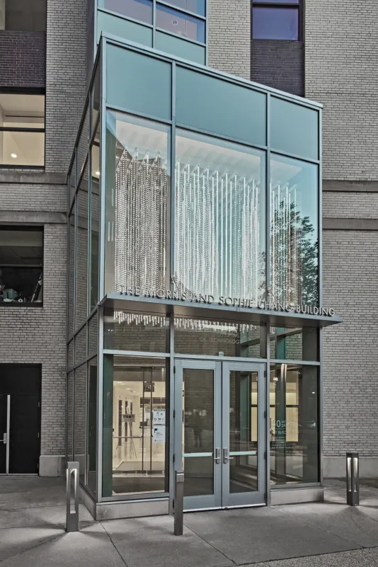 Glass entryway to a building with a diamond like sculptural work suspended in the doorway seen through the exterior glass. 
