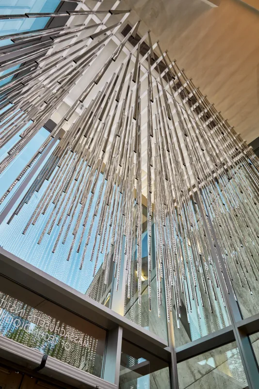 View from below of a sculptural work made of stainless steel rods and LED lights suspended in a glass entryway from the ceiling. 