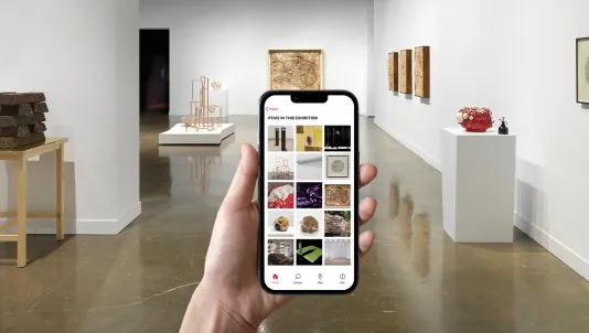Gallery with various sculptures in the background and a hand holding a phone with a screen that displays a collage of installation photography.