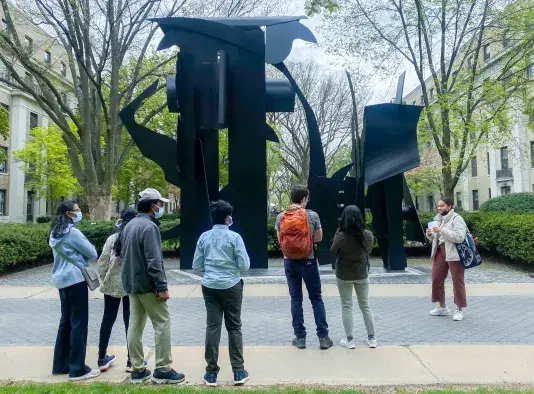 A small tour group stands facing a large, black steel sculpture with a tour guide on the right speaking to the group.
