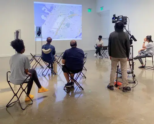 A small audience sits on gallery chairs in a gallery with a projector at the front and a speaker sitting to the right of the projector. A videographer stands to the right recording the lecture.