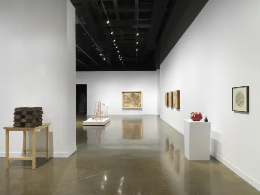Soil ingots are stacked on a wooden table. Beyond a floating wall is a lattice-like sculpture, a hanging rectangular sculpture, three smaller rectangular hanging reliefs, a red ceramic sculpture on pedestal and circular print.