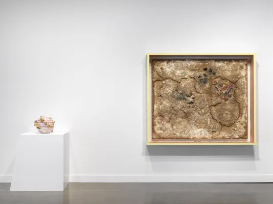 An orange and white striped beach ball with mushrooms growing on the sides sits on a geometric white pedestal sits to the left of a crusty framed artwork made of various materials resembling dried dirt. 