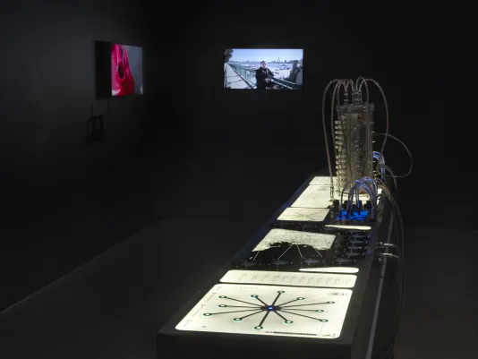 : In a dark gallery, a table with various controls is lit to display documents and scientific materials. Two adjacent monitors playing video glow in the background. 