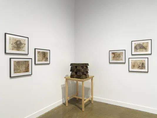 Stack of soil ingots on a light wooden table in a corner is surrounded on either side by clusters of three prints.