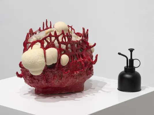 Sculpture of a lion’s mane mushroom that resembles a white foamy substance and grows inside a bright red ceramic vessel with a loose woven composition and many openings sits on a pedestal next to a matte black plant sprayer. 