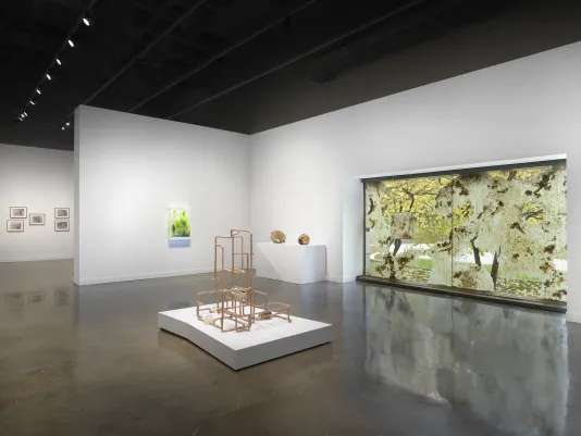 A beige lattice-like sculpture on a low white pedestal centers the gallery. A cluster of prints, a glowing green artwork, two small spherical sculptures, and a waxy window installation are in the background.