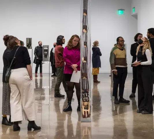 A group of gallery attendees stand around a large scale vertical sculpture that is encased in resin.