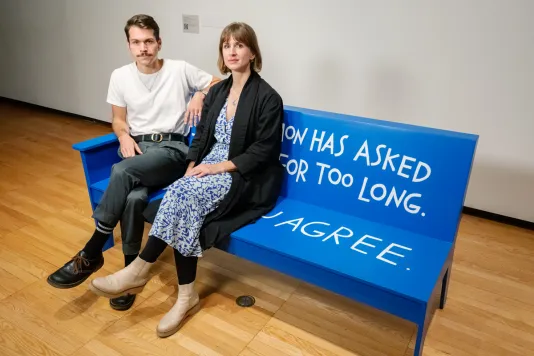 Two white people sit on a blue bench. On the left, Christopher Robert Jones sits and on the right Liza Sylvestre sits; both with legs crossed