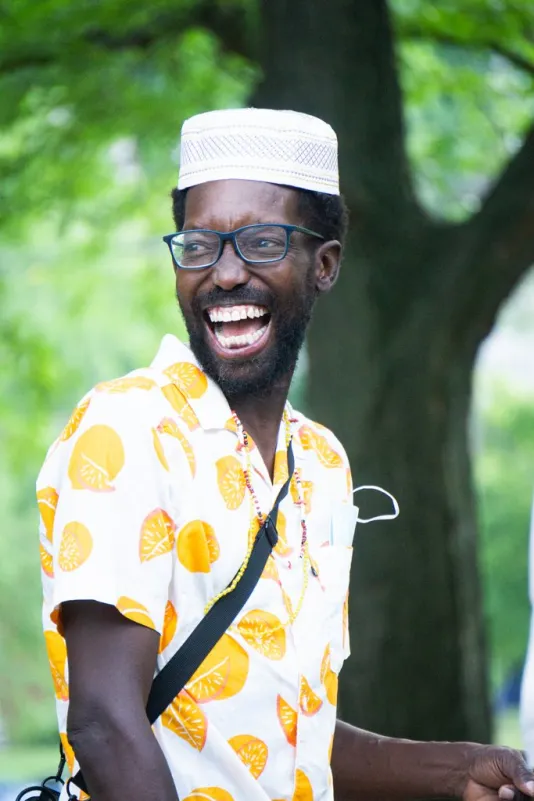 Standing outside, a dark skinned man with a full beard, smiles wide. He wears glasses, a white colored shirt adorned with oranges and a white hat