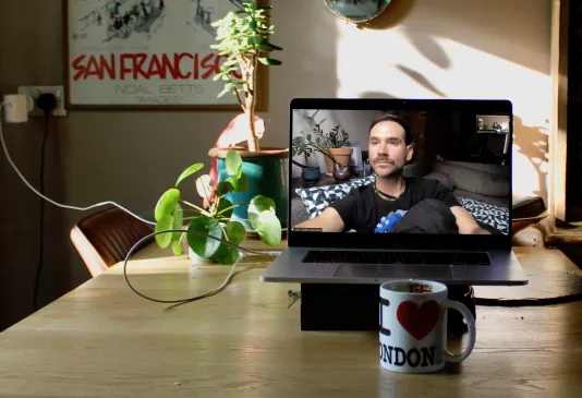 A macbook sits on a kitchen counter and displays zoom screen with a white person with short brown hair sitting on their couch.