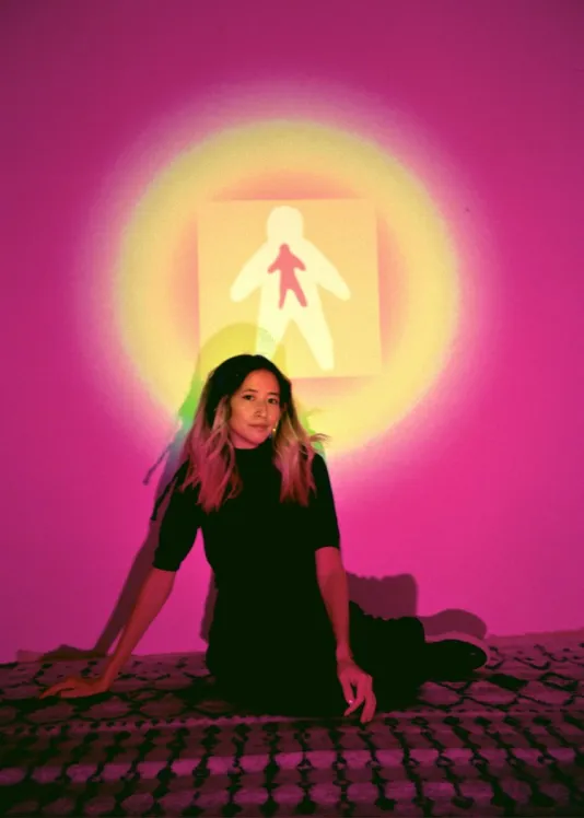 Wearing all black, Yo-Yo sits on a geometric carpet, in a room filled with magenta light. She has long bleach blonde hair with dark roots. A round projection of a humanlike figure beams on the wall behind her and illuminates her face.