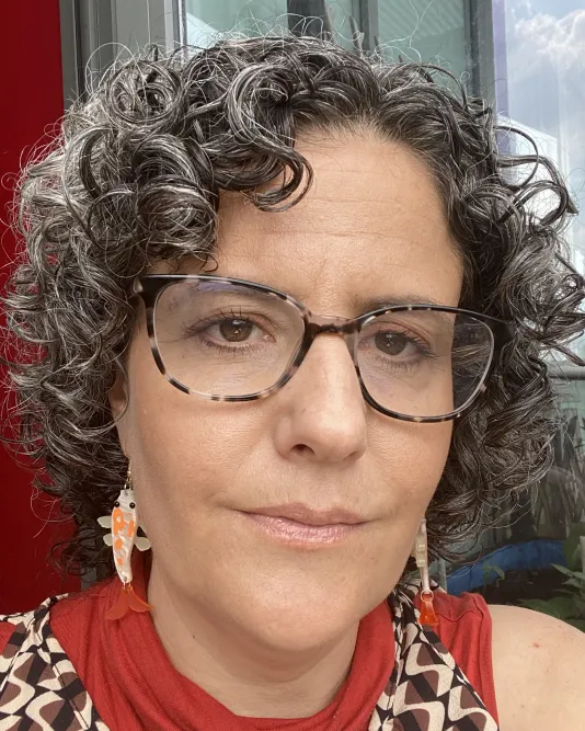 Headshot a woman with a neutral expression. She wears dark glasses and dangling earrings. She has dark curly hair with highlights of gray