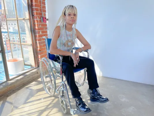 Person seated in a wheelchair amongst sculptural artwork in a gallery with floor to ceiling windows behind. Their head is turned to face the camera