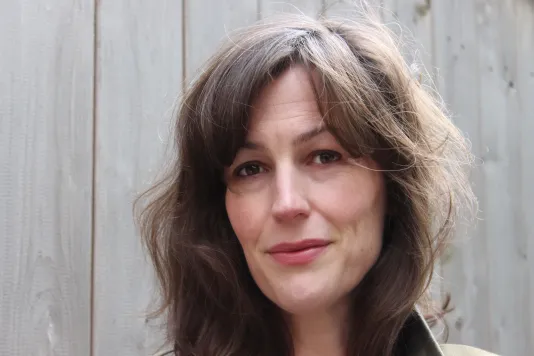 Close up photograph of a fair skinned woman, standing in front of a wooden wall. She has brown hair with bangs angling her face