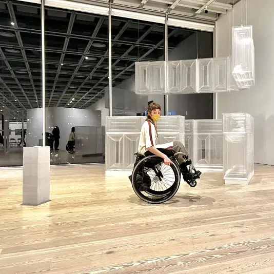 Person seated in a wheelchair amongst sculptural artwork in a gallery with floor to ceiling windows behind. Their head is turned to face the camera.