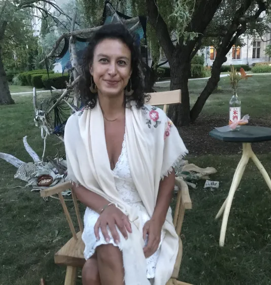 A brown-skinned woman with black curly hair smiles brightly, dressed all in white. She is seated outside in a hand-crafted chair