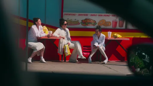 Three young women dressed in all white eat large bags of yellow chips and drink soda from brandless Styrofoam cups outdoors. Filmed from the driver’s seat of a car, a vent window and rear-view mirror frame the scene.