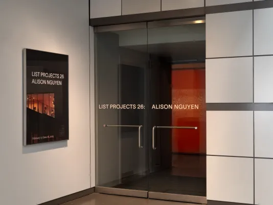 A dark poster that reads “LIST PROJECTS 26: ALISON NGUYEN” with image of orange glowing windows is hung next to two glass gallery doors with the same text and similar glowing light.  