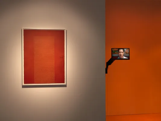 A white wall in the left foreground displays a reddish orange screen print featuring a rectangular block of yellow text framed in white. To the right is a video sculpture displaying an extreme close-up of a woman’s face. The walls in the background glow orange