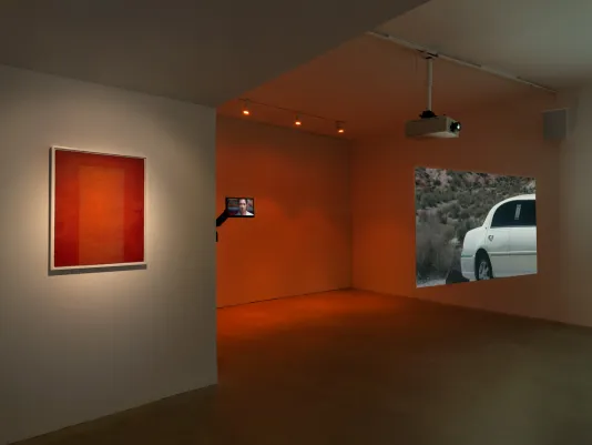 A burnt orange screen print is hung on the left wall. A screen held by a mount that looks like a car’s sideview mirror showing woman with black hair gazing at the viewer is hung off the wall to the right, lit with orange gallery lighting. To the far right, a projector shows the rear half of a white limousine against a desert landscape.