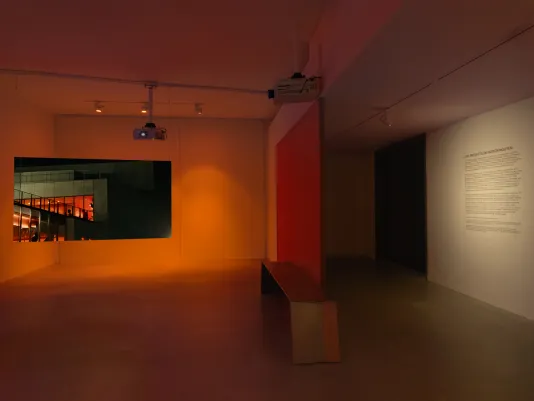 A 180° view of the gallery, to the left a large rectangular screen shows the exterior of a concrete building with a warm glow on the inside. The space is divided by a long aluminum bench and orange-red fabric wallwall text to the right.