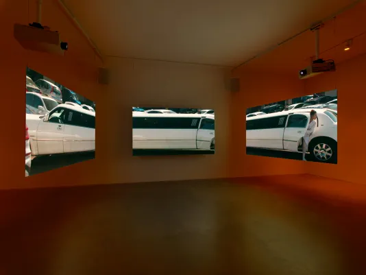 Three suspended screens show  a white stretch limousine in a desert landscape. The image of the limo is stretched across all three screens, in the portion at center, a suited figure appears to emerge from an open car door. 