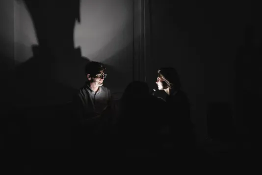 In a pitch black room, two people facing each other, hold up flashlights to illuminate their faces