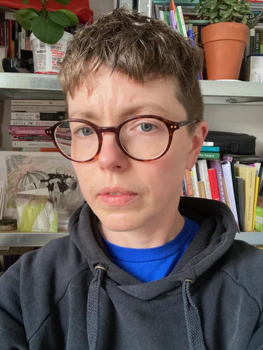 A person with fair skin, and short brown hair is pictured in front of shelves filled with colorful books and stacks of paper. They’re wearing a black hooded sweatshirt and round brown glasses.