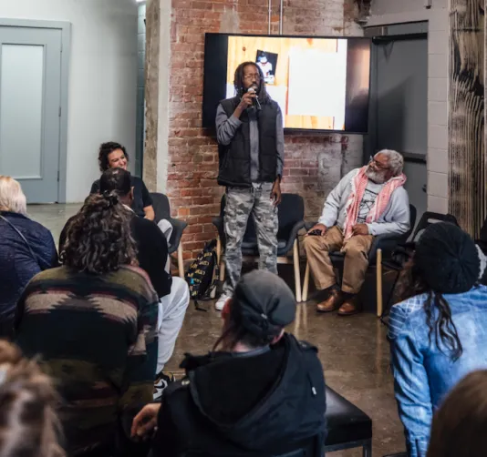 In front of a TV monitor, a black man with locks stands holding a microphone. He’s surrounded by a small crowd of people, all seated in chairs