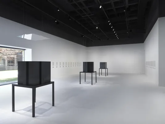  Three dark steel tables and geometric sculptures are surrounded by artworks in white frames hung horizontally in a white walled and floored gallery.