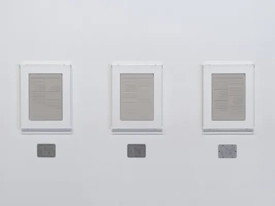 Three rectangular plaster works in white frames are hung side-by-side, each with a horizontal steel plaque below.
