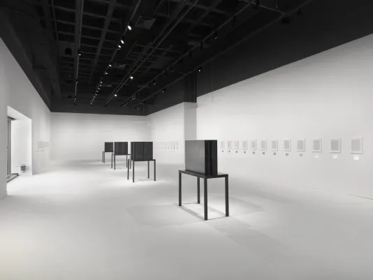 Four dark steel tables and geometric sculptures are surrounded by artworks in white frames hung horizontally in a white walled and floored gallery.