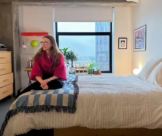A woman wearing a red sweater sits in a bedroom on a white bed in front of a window with two framed artworks to the right of the window.