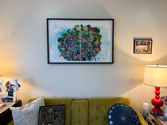 A black framed painting hangs on the wall above a green couch that is in between two lamps on end tables to the right and left of the couch. Only the top of the couch is visible at the bottom of the image.