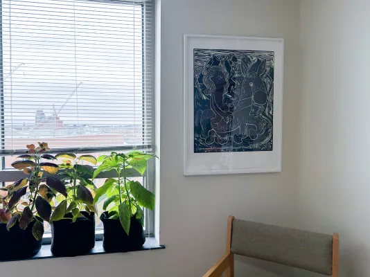 Dark artwork mounted in a white from on to a wall to the right of a window with plants on the windowsill.