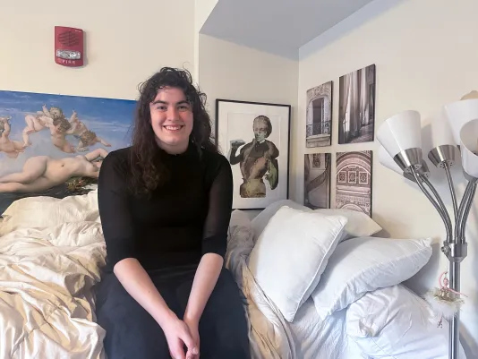 A woman dressed in black sits on a white bed to the left of a silver floor lamp. A large work of art is hung on the wall behind her, in addition to a smaller black framed artwork and four black and white images hung in a group farthest to the right.