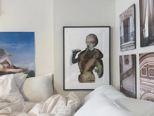 A black framed artwork is hung on the wall with four black and white images to the right hung in a grouping and part of another work of art is in view on the left. A white bed partly comes into view beneath the artworks.