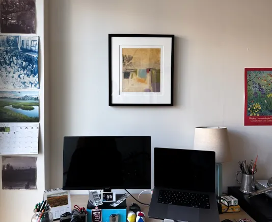 A black framed work of art is in the center of the image, hanging on the wall above a desk. Only part of the desk is in view and two computer screens can be seen sitting on the desk.