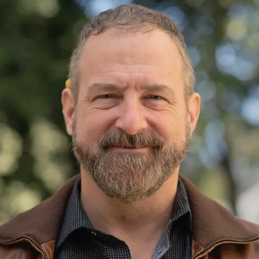 An image of Darrin Martin, a grinning, bearded, middle-aged, white man with light brown and grey speckled hair looking into the camera lens. He has a short haircut and wears a black button up shirt with a grid of small grey dots covered in part by a brown leather jacket opened at the front. The background is blurred trees with dapples of blue sky showing through.